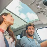 Pareja sonriente en un coche con techo panorámico. Ella, una conductora novel, disfruta del viaje.