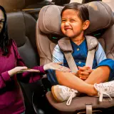 Una mujer sonríe mientras señala a un niño feliz sentado en su silla de coche. ¡Los niños en el coche están seguros y contentos!