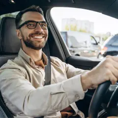 Hombre sonriente al volante, con gafas y camisa clara. Viaja en coche, quizás de camino a una cita importante, con la tranquilidad que ofrece la seguridad de Unicaja.