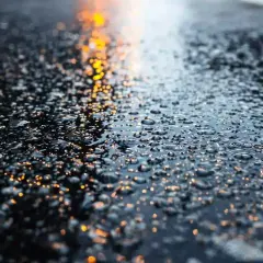 Gotas de agua brillan sobre la carretera mojada, reflejando luces anaranjadas.