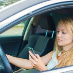 Mujer rubia al volante, mirando su móvil. Es importante evitar distracciones al conducir para mayor seguridad.