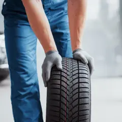 Man con guantes grises sostiene un neumático nuevo, listo para mejorar la adherencia del coche.