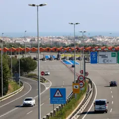 Peaje en la autopista Pau Casals con varios carriles, señales de tráfico y vehículos circulando. Al fondo, un paisaje urbano y el mar.
