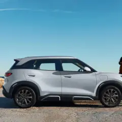 Pareja junto a un moderno coche híbrido blanco, con el mar y rocas al fondo.