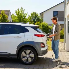 Hombre sonriente conecta su coche eléctrico a un cargador en casa. Conoce las tarifas del cargador de coche eléctrico para ahorrar.