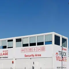 Edificio blanco con letreros "TRUCK VILLAGE" y "Security Area". Un poste con cámaras y luz ilumina el aparcamiento de camiones.