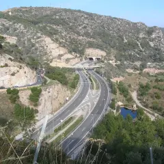 La autopista C32 serpentea entre montañas rocosas y vegetación, con un túnel y un puente.