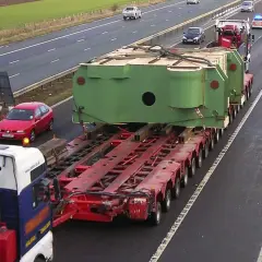 Un camión transporta una enorme pieza verde sobre una plataforma de múltiples ruedas. Un coche rojo y vehículos policiales acompañan este convoy de transportes especiales en la autopista.