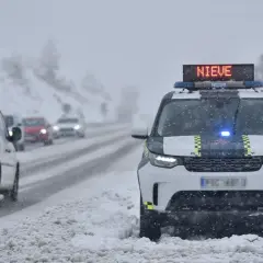 DGT alerta de nieve en carreteras. Un coche patrulla con luces azules y el mensaje "NIEVE" en su panel luminoso se detiene en un arcén nevado.