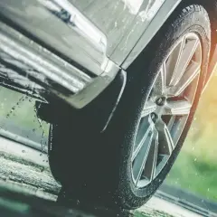 Rueda de coche sobre asfalto mojado, con gotas de agua salpicando, indicando una posible **pérdida de adherencia** en condiciones de lluvia.