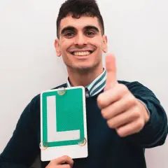 Joven sonriente con letrero "L" y pulgar arriba, listo para ser novel al volante.