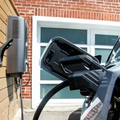Cargador bidireccional V2H conectado a un coche eléctrico. La unidad de carga está montada en una pared de madera, con una casa de ladrillo de fondo.