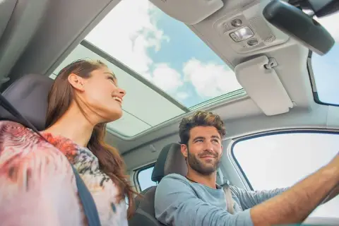 Pareja sonriente en un coche con techo panorámico. Ella, una conductora novel, disfruta del viaje.
