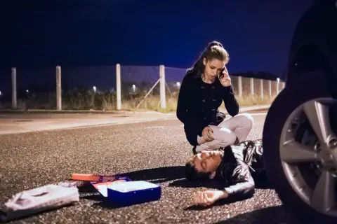 Una mujer llama a emergencias junto a un hombre tendido en la carretera. A su lado, un botiquín abierto. ¿Qué significa pas? ¿Una vida en peligro?