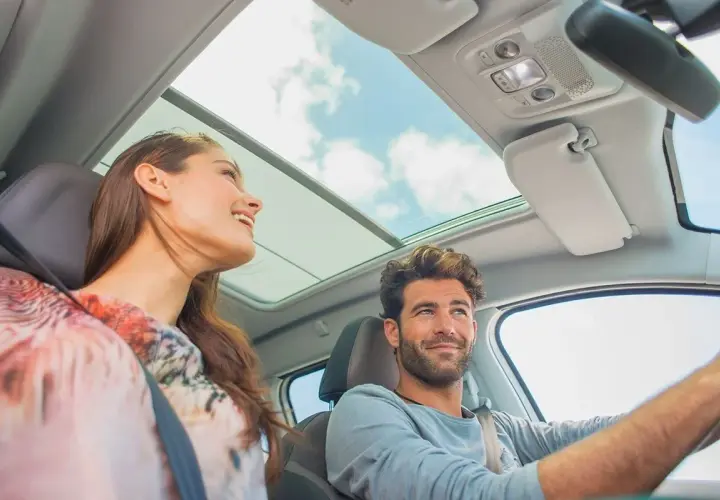Pareja sonriente en un coche con techo panorámico. Ella, una conductora novel, disfruta del viaje.