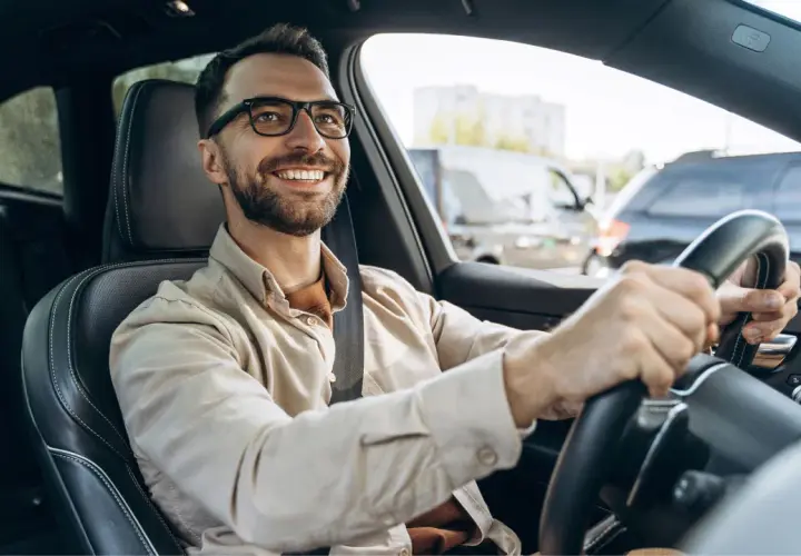 Hombre sonriente al volante, con gafas y camisa clara. Viaja en coche, quizás de camino a una cita importante, con la tranquilidad que ofrece la seguridad de Unicaja.