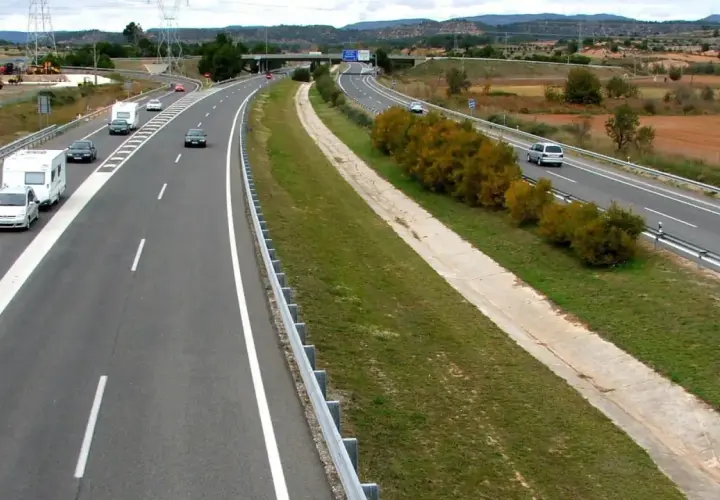 Autopista con varios coches y caravanas. Se aprecian las áreas de servicio AP2 al fondo, con vegetación y campos.