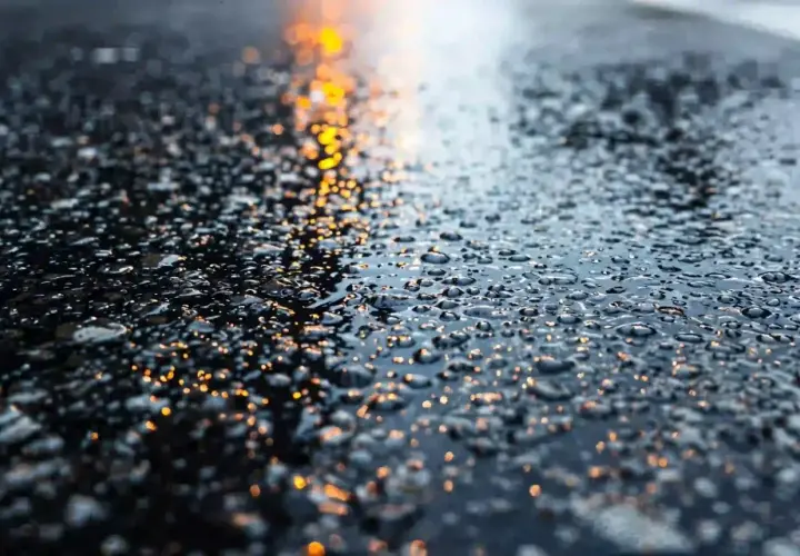 Gotas de agua brillan sobre la carretera mojada, reflejando luces anaranjadas.
