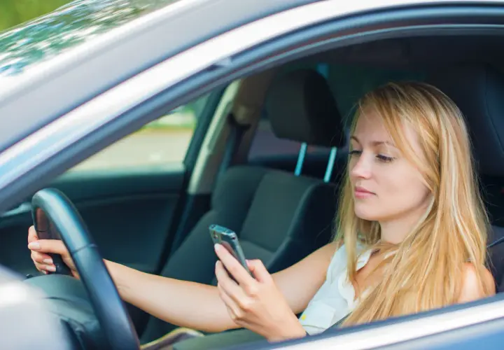 Mujer rubia al volante, mirando su móvil. Es importante evitar distracciones al conducir para mayor seguridad.
