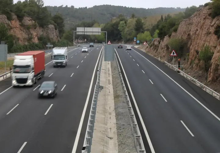 Tráfico en las autopistes catalanes, con camiones y coches circulando por una carretera de varios carriles rodeada de vegetación y rocas.
