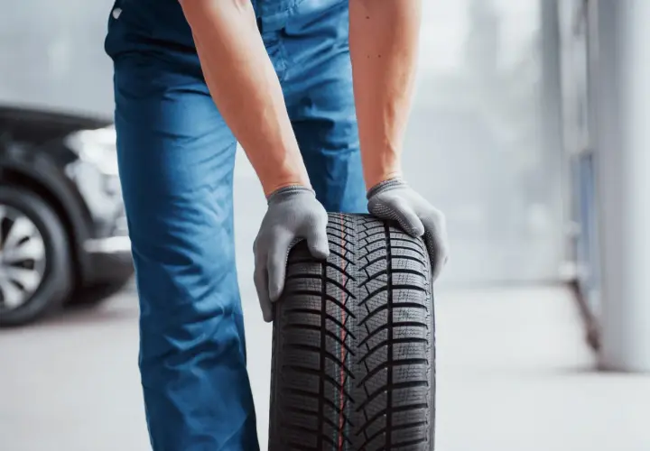 Man con guantes grises sostiene un neumático nuevo, listo para mejorar la adherencia del coche.