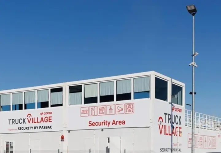 Edificio blanco con letreros "TRUCK VILLAGE" y "Security Area". Un poste con cámaras y luz ilumina el aparcamiento de camiones.
