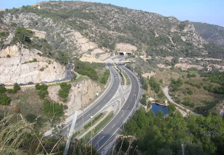 La autopista C32 serpentea entre montañas rocosas y vegetación, con un túnel y un puente.