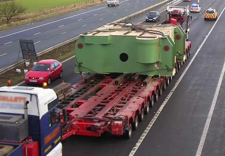 Un camión transporta una enorme pieza verde sobre una plataforma de múltiples ruedas. Un coche rojo y vehículos policiales acompañan este convoy de transportes especiales en la autopista.