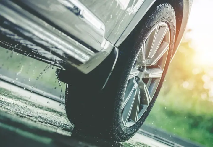 Rueda de coche sobre asfalto mojado, con gotas de agua salpicando, indicando una posible **pérdida de adherencia** en condiciones de lluvia.