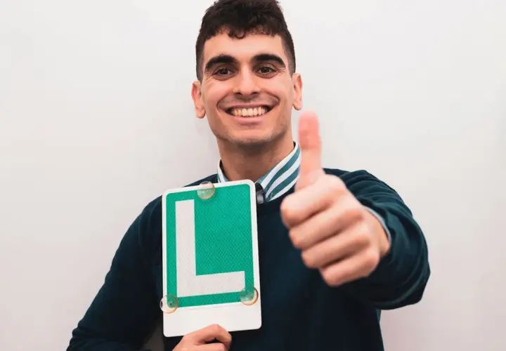 Joven sonriente con letrero "L" y pulgar arriba, listo para ser novel al volante.