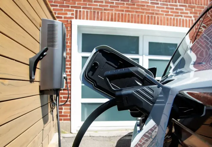 Cargador bidireccional V2H conectado a un coche eléctrico. La unidad de carga está montada en una pared de madera, con una casa de ladrillo de fondo.