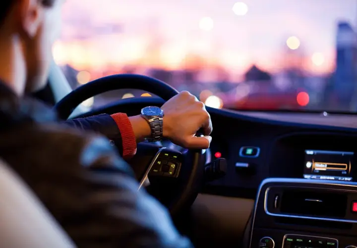 Hombre con reloj conduce a mayor velocidad, con luces de ciudad borrosas al atardecer.