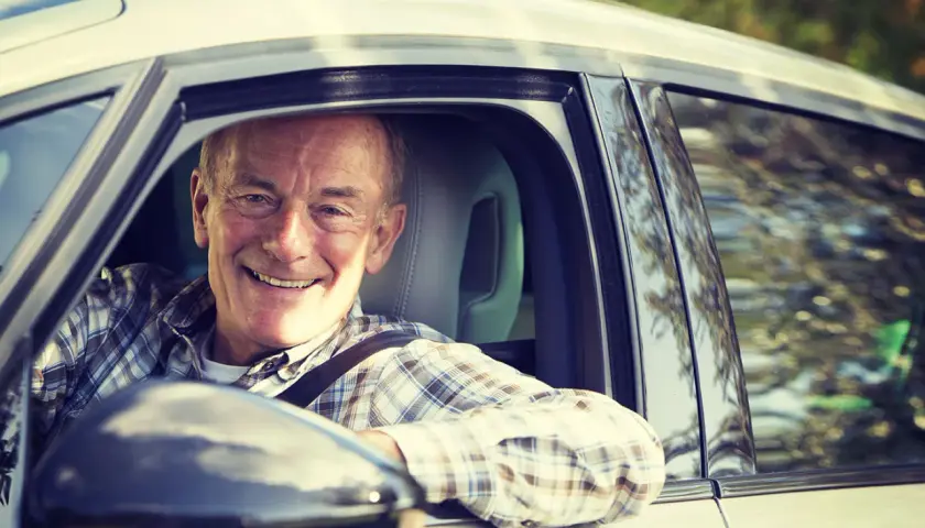 Hombre mayor sonriente al volante. Las personas mayores suelen tener menos accidentes en situaciones de conducción tranquila y familiar.