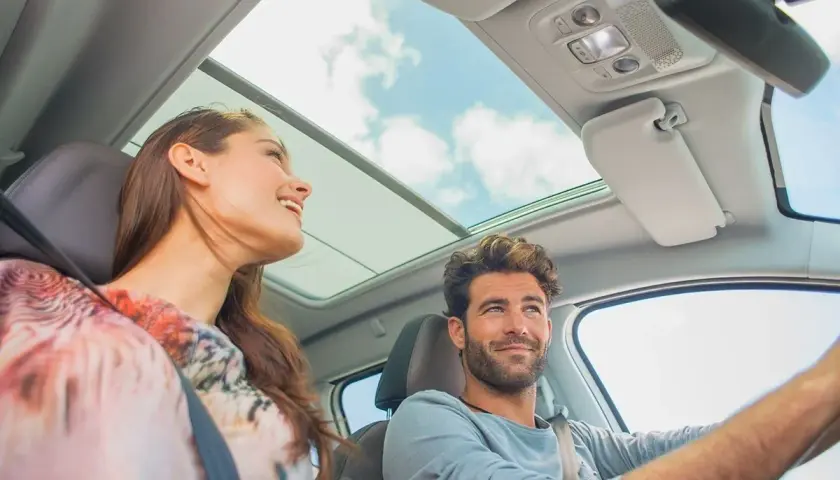 Pareja sonriente en un coche con techo panorámico. Ella, una conductora novel, disfruta del viaje.