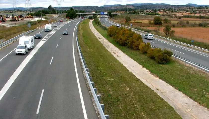 Autopista con varios coches y caravanas. Se aprecian las áreas de servicio AP2 al fondo, con vegetación y campos.