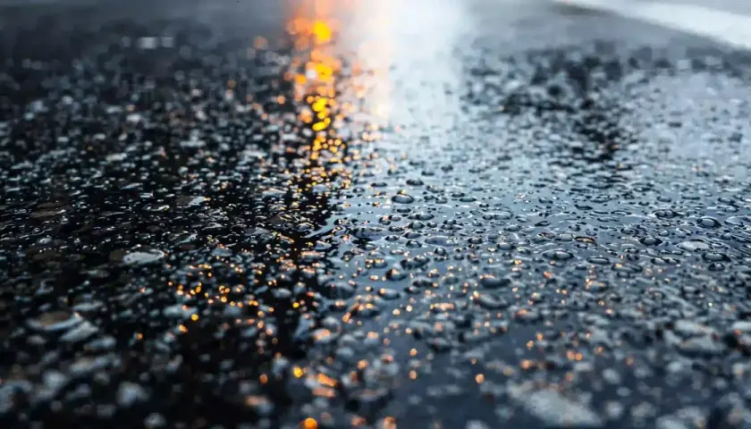 Gotas de agua brillan sobre la carretera mojada, reflejando luces anaranjadas.