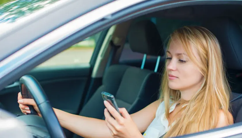 Mujer rubia al volante, mirando su móvil. Es importante evitar distracciones al conducir para mayor seguridad.