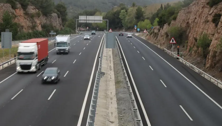 Tráfico en las autopistes catalanes, con camiones y coches circulando por una carretera de varios carriles rodeada de vegetación y rocas.