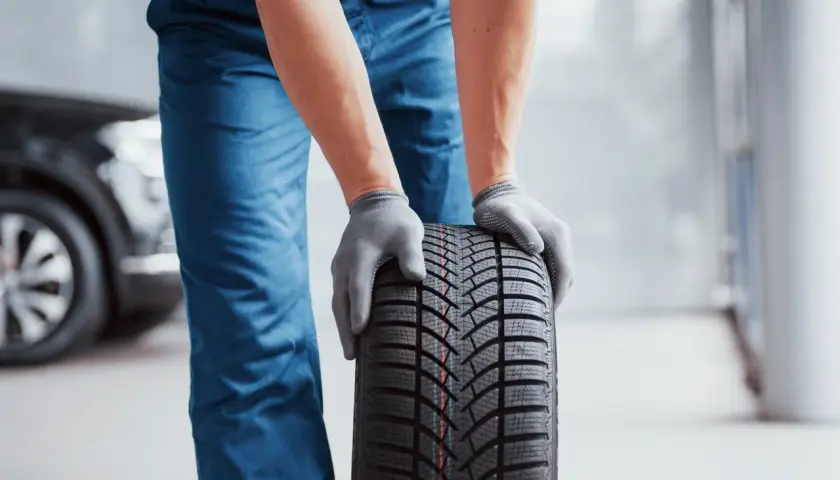Man con guantes grises sostiene un neumático nuevo, listo para mejorar la adherencia del coche.