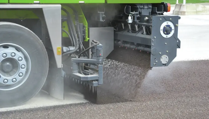 Máquina esparciendo material granular sobre el asfalto, parte de un proceso de reciclado.