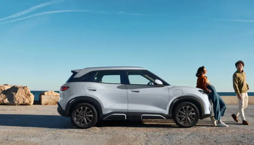 Pareja junto a un moderno coche híbrido blanco, con el mar y rocas al fondo.