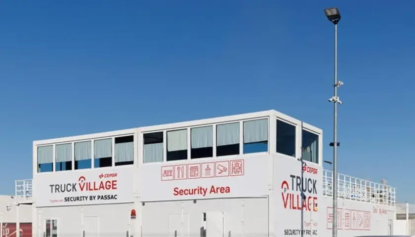 Edificio blanco con letreros "TRUCK VILLAGE" y "Security Area". Un poste con cámaras y luz ilumina el aparcamiento de camiones.