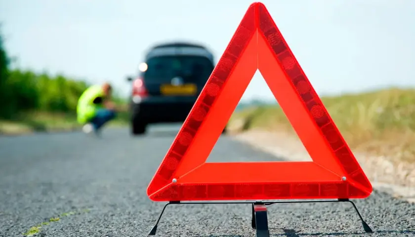 Triángulo de señalización de emergencia en carretera. Un coche averiado y una persona con chaleco reflectante en el fondo. Esquema pas.