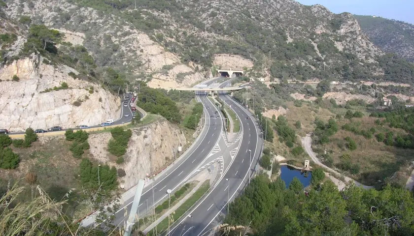 La autopista C32 serpentea entre montañas rocosas y vegetación, con un túnel y un puente.