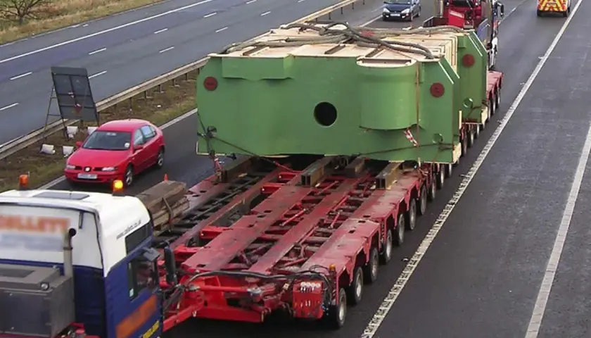 Un camión transporta una enorme pieza verde sobre una plataforma de múltiples ruedas. Un coche rojo y vehículos policiales acompañan este convoy de transportes especiales en la autopista.
