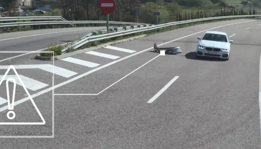 Un coche blanco se detiene cerca de un parachoques dañado en la carretera, indicando un accidente por objeto en la carretera.