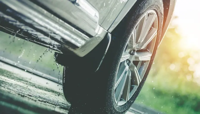 Rueda de coche sobre asfalto mojado, con gotas de agua salpicando, indicando una posible **pérdida de adherencia** en condiciones de lluvia.