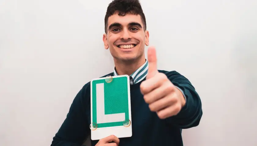 Joven sonriente con letrero "L" y pulgar arriba, listo para ser novel al volante.