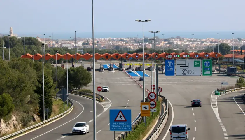 Carriles de peaje con barreras y señales de tráfico. Un coche blanco se acerca al peaje garraf, con la ciudad y el mar al fondo.