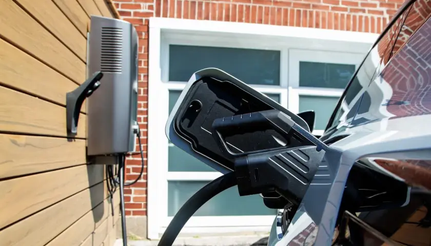 Cargador bidireccional V2H conectado a un coche eléctrico. La unidad de carga está montada en una pared de madera, con una casa de ladrillo de fondo.